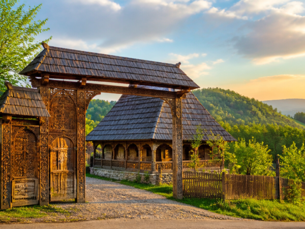 A stunning panoramic view of a traditional wooden house in Maramureș, Romania, featuring a tall carved wooden gate with intricate symbolic motifs, a steep wooden shingled roof, surrounded by lush green rolling hills in soft spring light. Photorealistic style, high detail architectural photography, warm natural lighting, golden hour, vibrant colors, traditional rural Eastern European village atmosphere, 8K resolution.